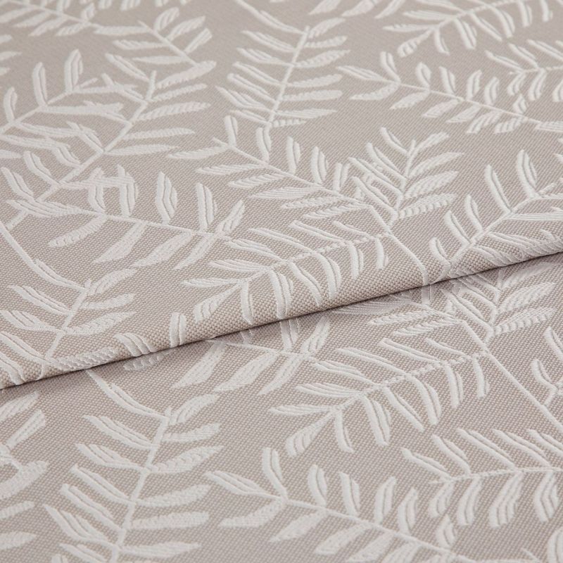 Beige jacquard fabric with raised white fern motif; folded and overlapped; close-up of an upholstery/curtain sample on a flat surface under soft, neutral lighting.