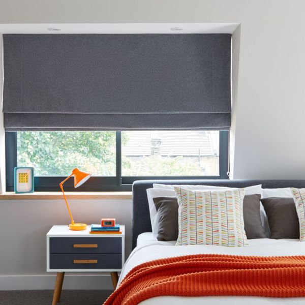 Grey fabric Roman blind lowered and mostly closed, filtering light with a small gap; bedroom interior with bed, patterned cushions, orange throw and bedside table with lamp.
12:01