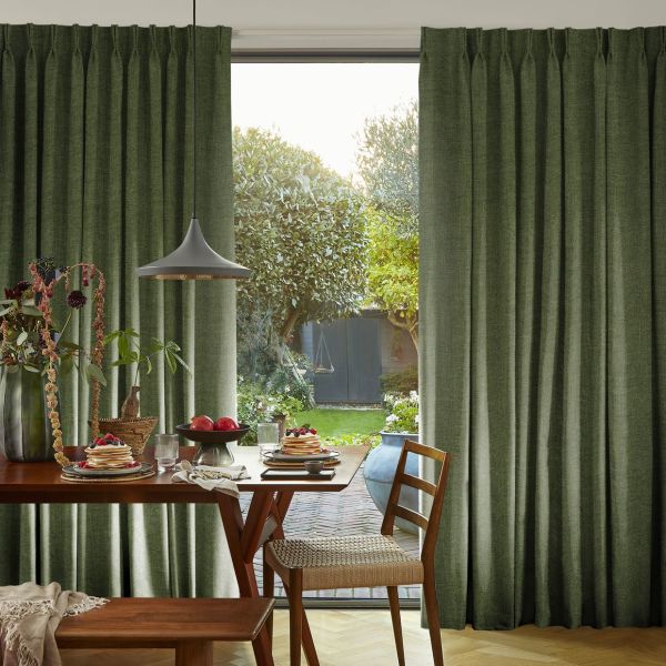 Green pleated floor-length curtains parted to reveal a glass sliding door, filtering soft daylight. Dining table set with pancakes and fruit, pendant lamp overhead, backyard garden and trees visible.
