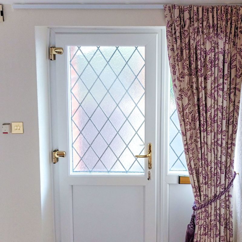 Floral pleated curtains, drawn right and tied back with matching rope, partially covering a white diamond-leaded glass door with brass handle; soft daylight filters into the entry hall.