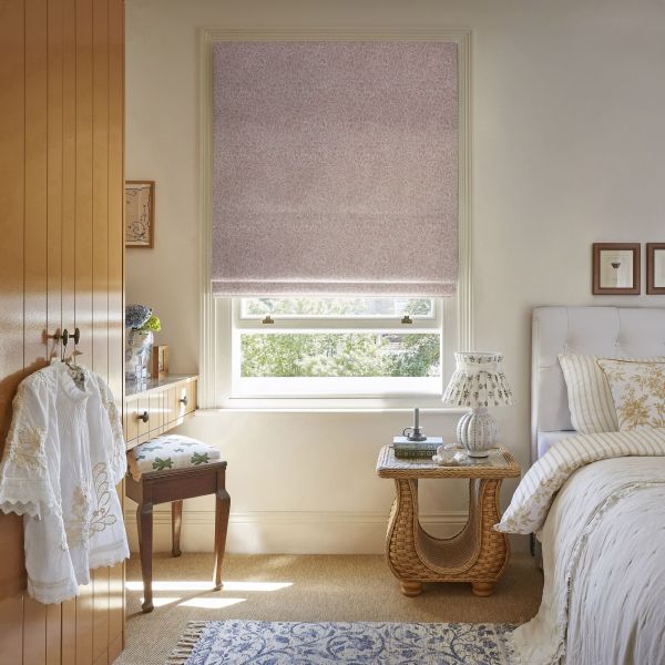 Pink patterned Roman blind, partially lowered over a sash window, softens incoming sunlight; bedroom with upholstered bed, wicker side table and lamp, wooden wardrobe and leafy outside view.