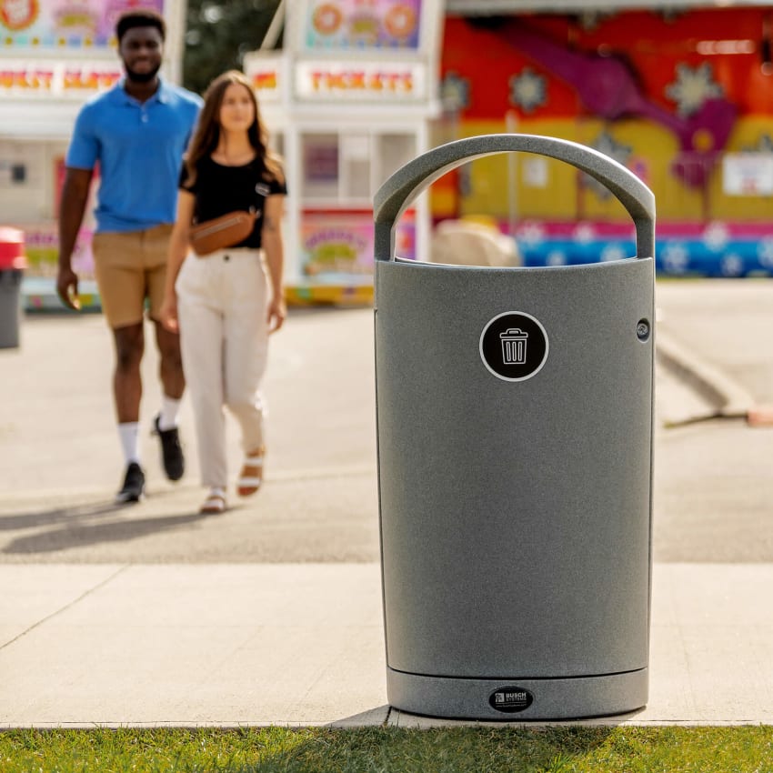 single grey waste bin outdoors at fall fair with midway rides and people walking by