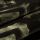 Velvet-like patterned fabric with dark olive and black abstract geometric print; folded into overlapping layers, showing soft nap and woven texture in dim, close-up studio lighting.