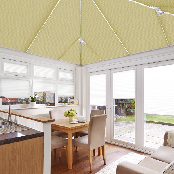 Honey-yellow pleated roof blinds and white pleated window/door blinds covering a conservatory's pitched ceiling and frames, filtering daylight over a dining table, chairs, kitchen sink and garden patio.