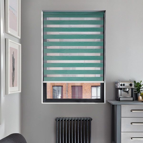 Teal zebra roller blind, lowered mid-window, with alternating opaque and sheer horizontal bands filtering daylight; set in a gray kitchen room with radiator beneath, countertop espresso machine and cactus prints.