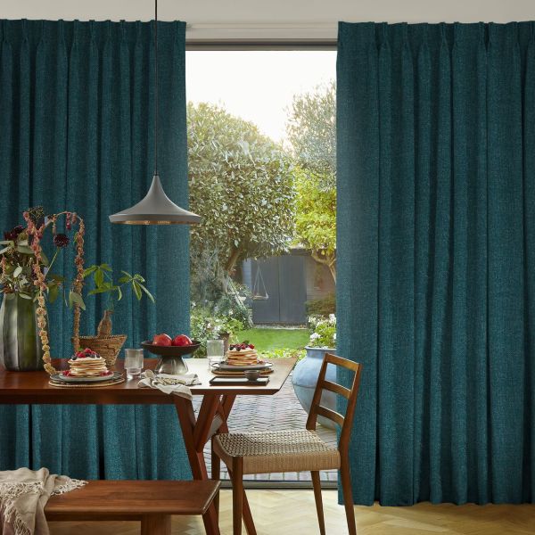 Teal pleated full-length curtains, slightly parted; partially open, filtering daylight and framing a glass door. Dining table set for breakfast with wooden chairs; view into a garden and shed.