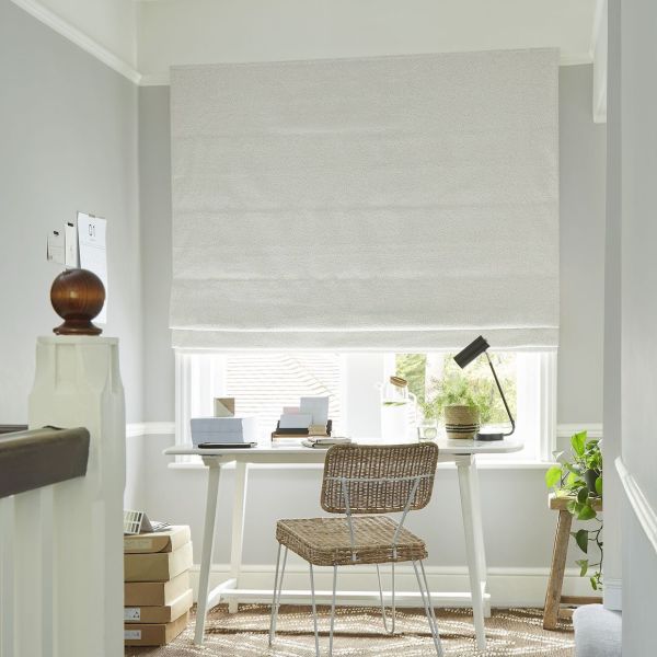 White textured Roman shade lowered over a window, blocking most light and letting soft daylight from the bottom; small home office desk with wicker chair, lamp and potted plants. 01