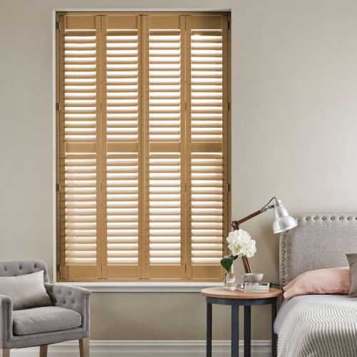Wooden plantation shutters covering a tall recessed window, full-height four-panel design with adjustable slats; shutters closed and diffusing warm daylight into a neutral, furnished bedroom with armchair and bedside table.