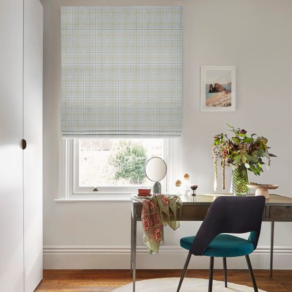 Grey plaid Roman blind lowered over a window, filtering daylight. In a bright home office: wooden desk with teal chair, mirror, cascading floral vase and framed coastal print, garden view.