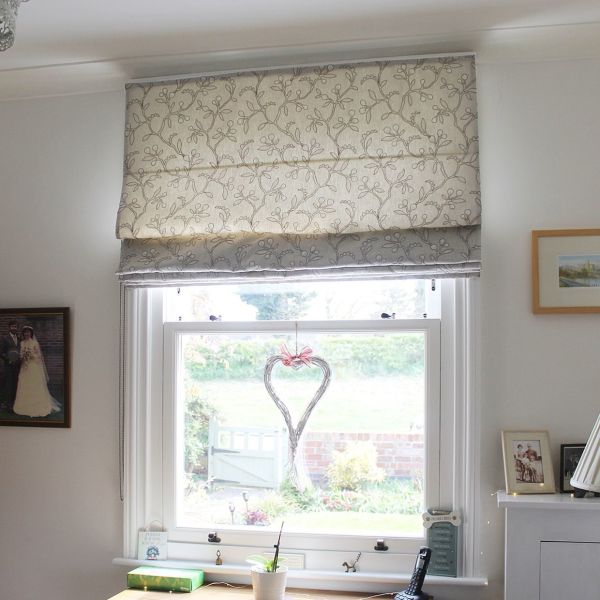 Cream fabric Roman blind with grey vine embroidery, lowered halfway and softly filtering daylight; hangs over a white-framed sash window in a bright room with photos, desk and garden view.
