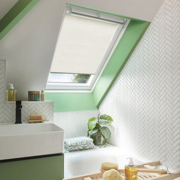 A beige pleated skylight blind, mounted inside an angled roof window, half-lowered, filtering daylight over a green-trimmed bathroom with white herringbone tiles, sink, bathtub, toiletries and a potted plant.