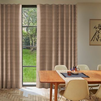 Patterned beige full-length curtains frame a narrow window, drawn closed with a central gap revealing a lawn and tree, in a lit dining room with wooden table and chairs.