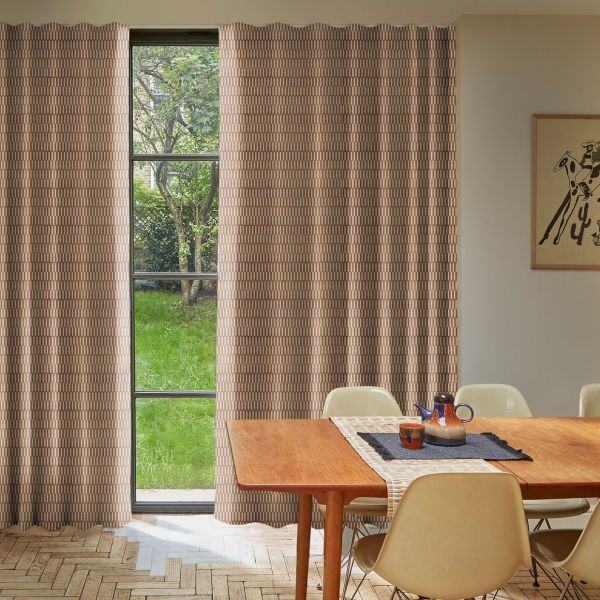 Patterned beige full-length curtains frame a narrow window, drawn closed with a central gap revealing a lawn and tree, in a lit dining room with wooden table and chairs.