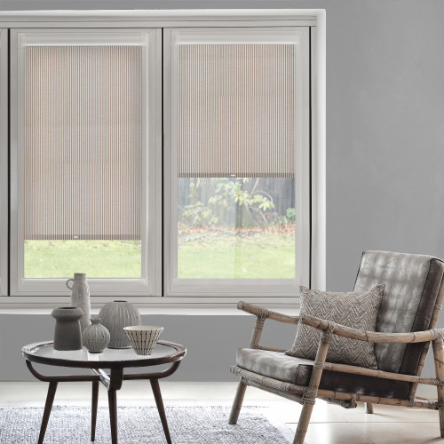 Striped beige pleated blinds cover the upper halves of twin windows, partially lowered and softly filtering daylight; modern living room with wooden-framed armchair, patterned cushion, small coffee table, lawn view.