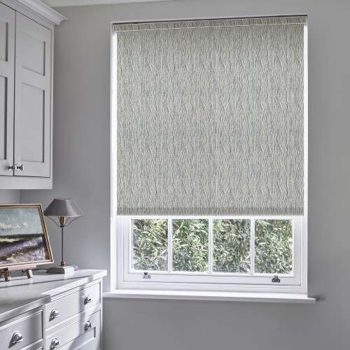 A patterned roller blind with subtle wavy grey lines, lowered to the sill, filtering daylight; in a grey room over a window above a dresser with lamp, garden visible.