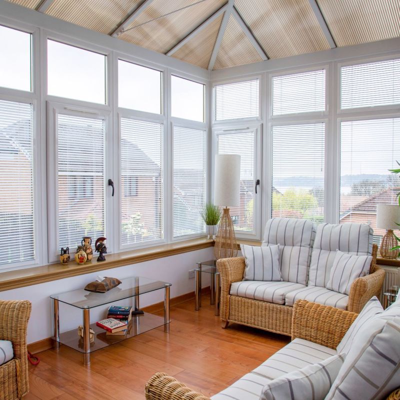 White horizontal blinds covering tall conservatory windows, slats partially tilted, filtering soft daylight into a sunroom with wicker sofas, striped cushions, wood floor, glass coffee table, and lamps.