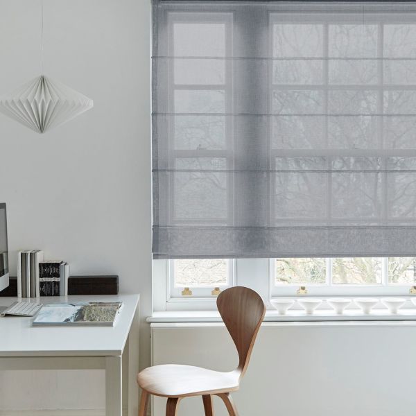 Sheer grey Roman blind, lowered to mid-window with soft horizontal folds, filtering diffused daylight; minimal home office with white desk, wooden chair, paper pendant lamp, and faint tree silhouettes outside.