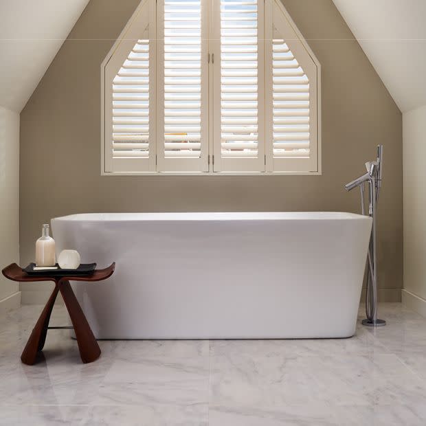 White plantation shutters cover a pitched-arched window, slats tilted to filter daylight; a freestanding white tub sits beneath with chrome floor-mounted faucet, marble floor, neutral sloped attic bathroom.