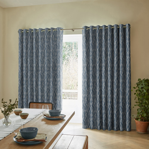 Blue patterned grommet-top curtains, hung on a metal rod, are partially drawn and filtering daylight through a central gap; dining room with wooden table settings, neutral walls, and a potted plant.