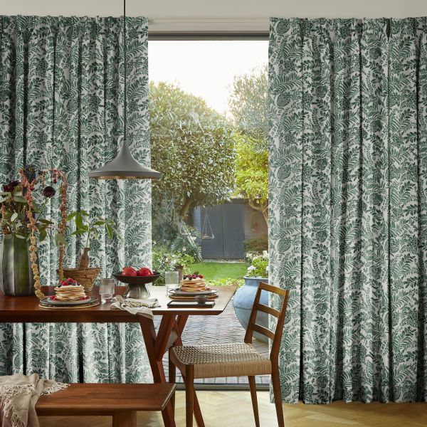 Botanical-print, floor-length curtains at a sliding glass door, parted to frame and let sunlight through; dining table with pancakes and vase beneath a pendant lamp opening onto a sunlit garden.