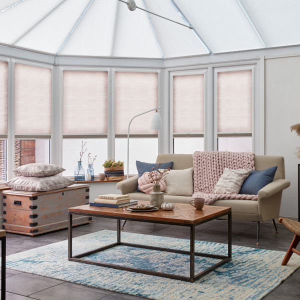 Pleated cellular blinds covering bay and roof windows, pale blush and fully closed, softly diffusing daylight; bright conservatory living area with beige sofa, coffee table, chunky pink throw and rug.