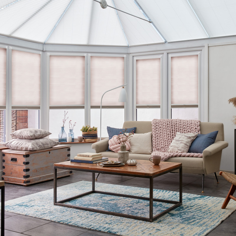 Pleated cellular blinds covering bay and roof windows, pale blush and fully closed, softly diffusing daylight; bright conservatory living area with beige sofa, coffee table, chunky pink throw and rug.