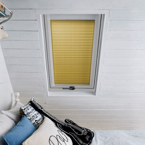 Yellow pleated blind pulled down over a rectangular skylight, filtering daylight; attic bedroom with white shiplap walls and a bed stacked with decorative pillows and a guitar-shaped cushion.