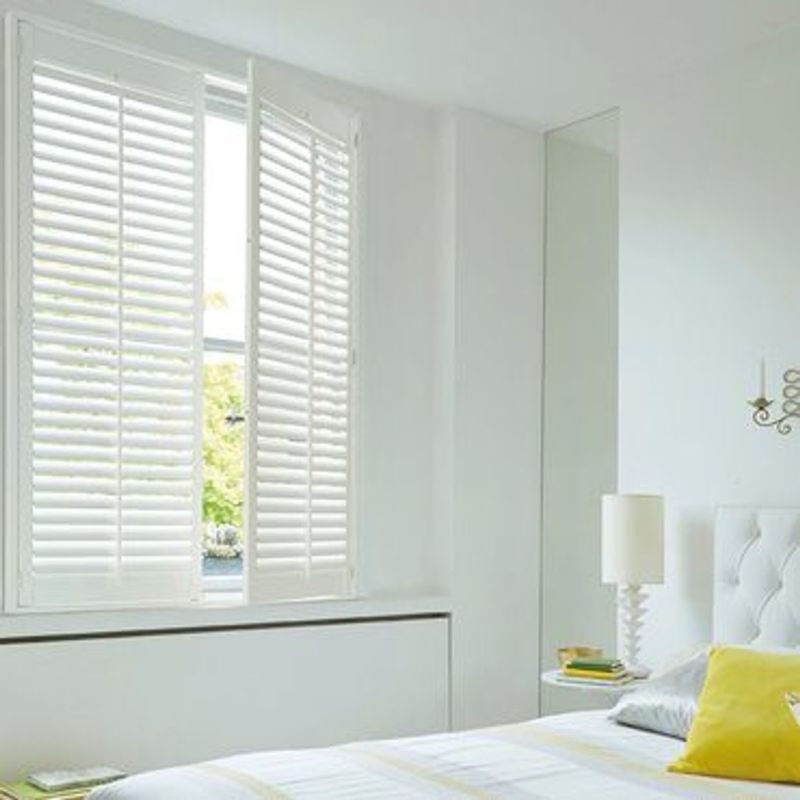 White plantation shutters on a three-panel window, slatted panels mostly closed with one panel ajar, filtering daylight into a minimalist white bedroom with tufted headboard, lamp and yellow pillow.