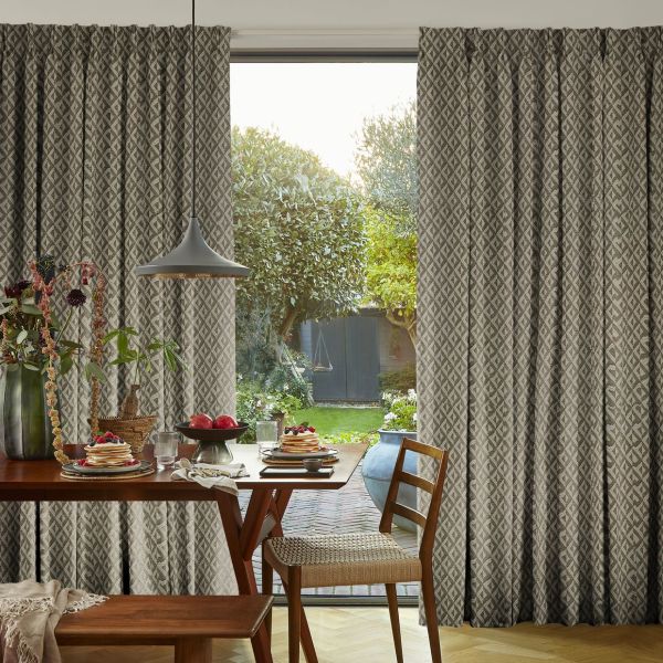 Patterned pleated floor-to-ceiling curtains with diamond motif, pulled aside to reveal a sliding glass door and sunlit backyard; wooden dining table set for breakfast, pendant lamp overhead, green garden view.