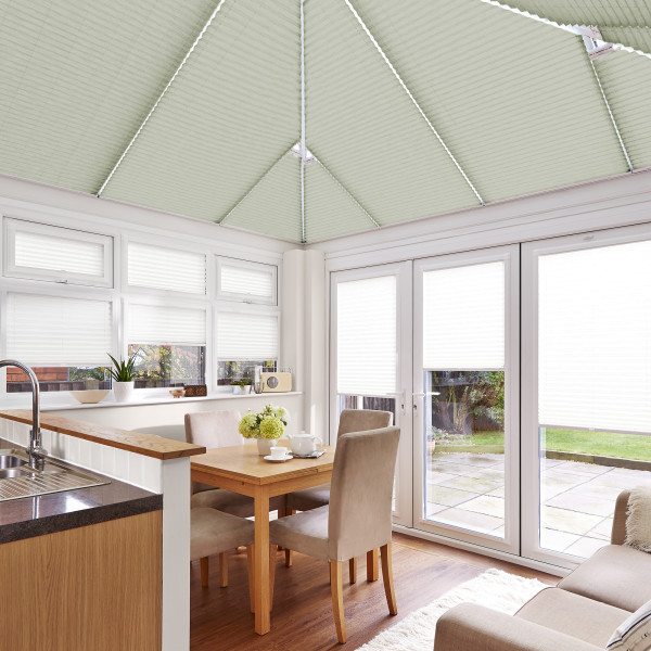 Pleated, pale-green roof blinds drawn across the pitched conservatory ceiling and white window and door blinds, filtering daylight in a bright dining-kitchen with wooden table, beige chairs and garden view.