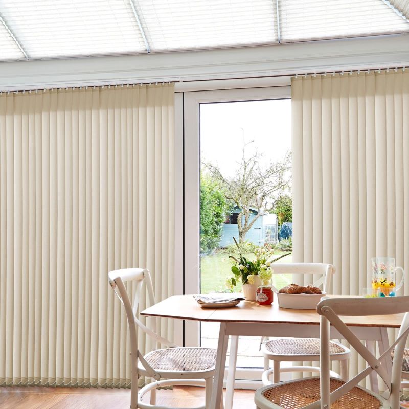 Beige vertical blinds (floor-to-ceiling) across glass patio doors; slats mostly closed, partially drawn revealing central gap, softly filtering daylight. Dining nook with wooden table, chairs, vase and garden view.