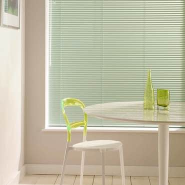 Horizontal pale-green Venetian blinds cover the window, lowered and closed, softly filtering daylight; a dining nook with a white round table, translucent lime-green chair, stool, and light wood floor.