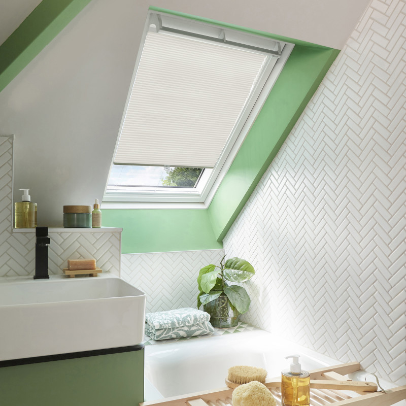 White pleated skylight blind, half-lowered and filtering soft daylight, covers a roof window above a sloped bathroom with mint-green trim, white herringbone tiles, sink, bathtub, plant and bath accessories.