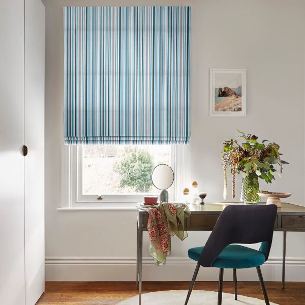 Blue-and-white striped Roman blind, lowered with neat folds, filtering soft daylight into a small home office featuring a wooden desk, teal chair and a vase of flowers.