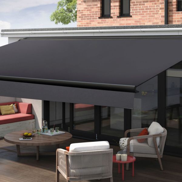 Dark retractable fabric awning with metal roller and valance — fully extended — shading a glass-walled patio furnished with cushioned armchairs, round wooden table and built-in bench beneath brick facade, daylight.