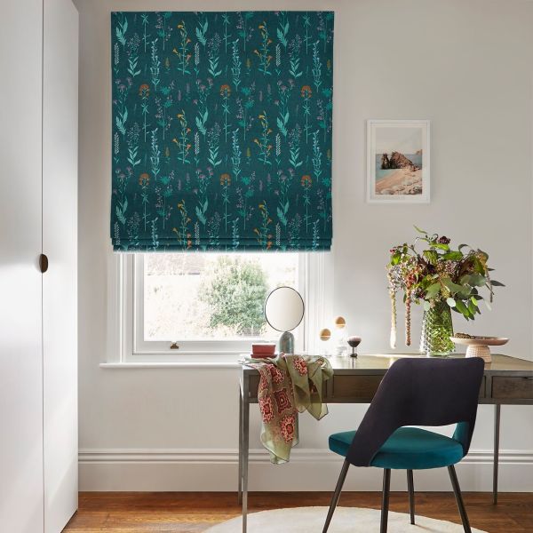 A teal, patterned Roman blind with vertical floral motifs, fully lowered and folded at the bottom, filtering daylight over a white window; small desk, chair, vase of flowers, neutral wall.
