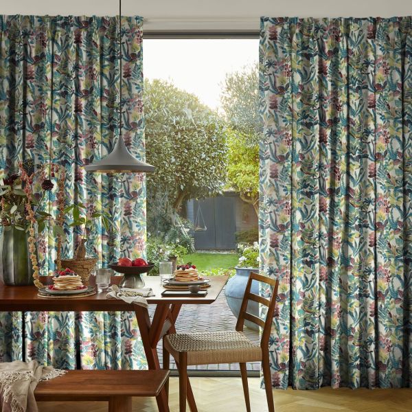 Pleated floor-length floral curtains (teal, pink, yellow) parted centrally, revealing a sliding glass door and admitting soft morning light; wooden breakfast table with pancakes, pendant lamp, woven chair, and garden.