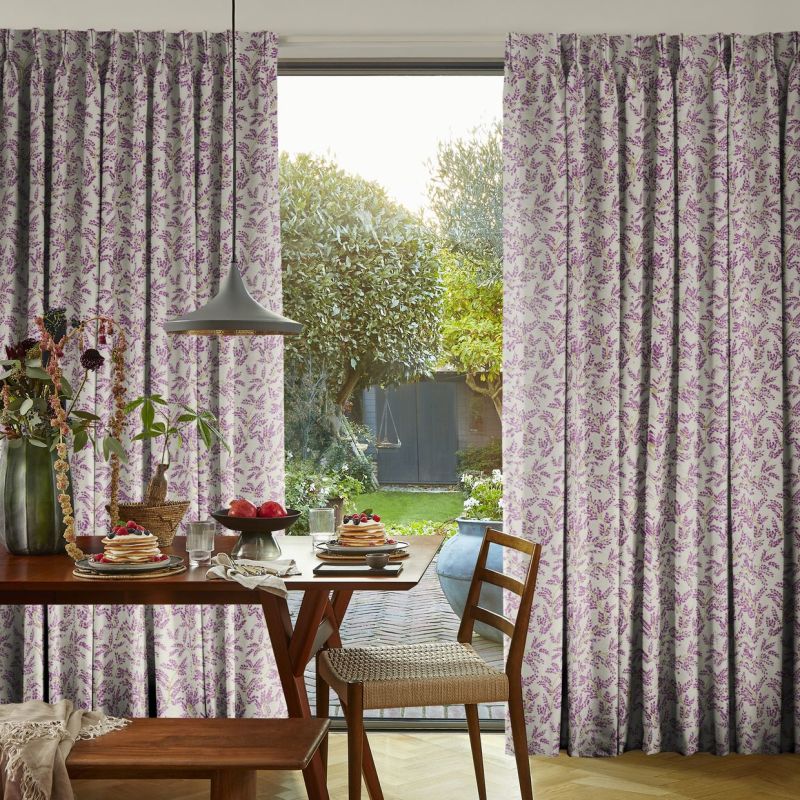Pleated, floor-length curtains with a purple sprig pattern, hung at a sliding door and drawn apart; framing a sunlit backyard view and a breakfast-set dining table under a pendant light.