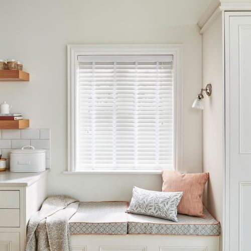 White horizontal slatted blinds, fully lowered and mostly closed, filtering soft daylight; above a built-in cushioned window seat with patterned cushions, a throw, wall sconce and floating shelves in a pale kitchen nook.

BREAD