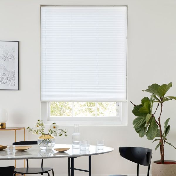 White pleated cellular blind, lowered nearly to the sill with horizontal seams, diffusing daylight; set in a white dining area with marble table, black chairs and a potted fiddle‑leaf fig.