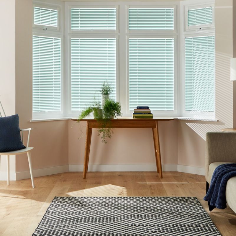 Horizontal white Venetian blinds cover a five-panel bay window, closed and filtering daylight; wooden console holds potted plants in a living room with wood floor, rug, chair and sofa.