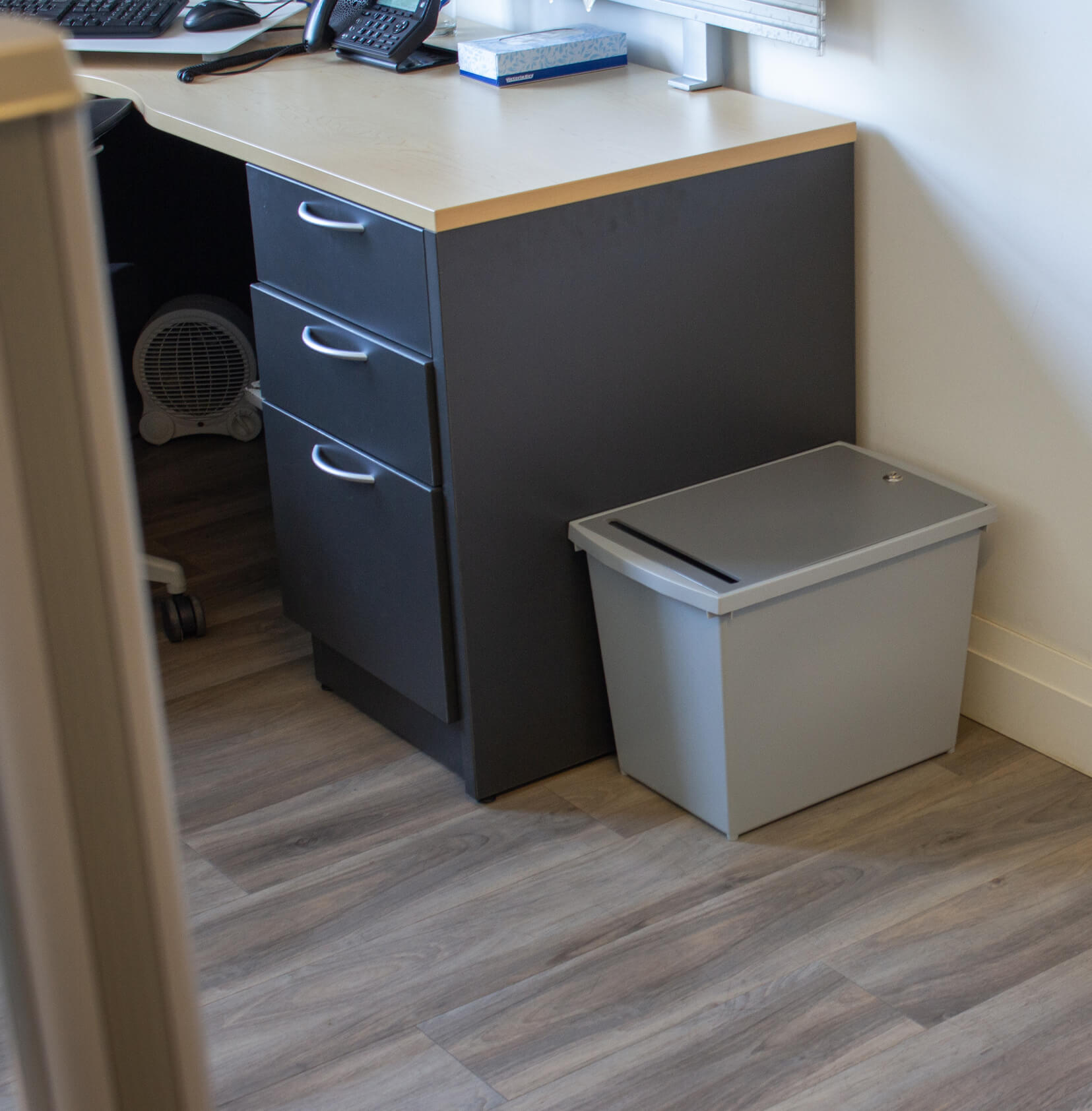 grey plastic personal document container for collecting important papers for shredding in an office sitting on the floor beside a desk