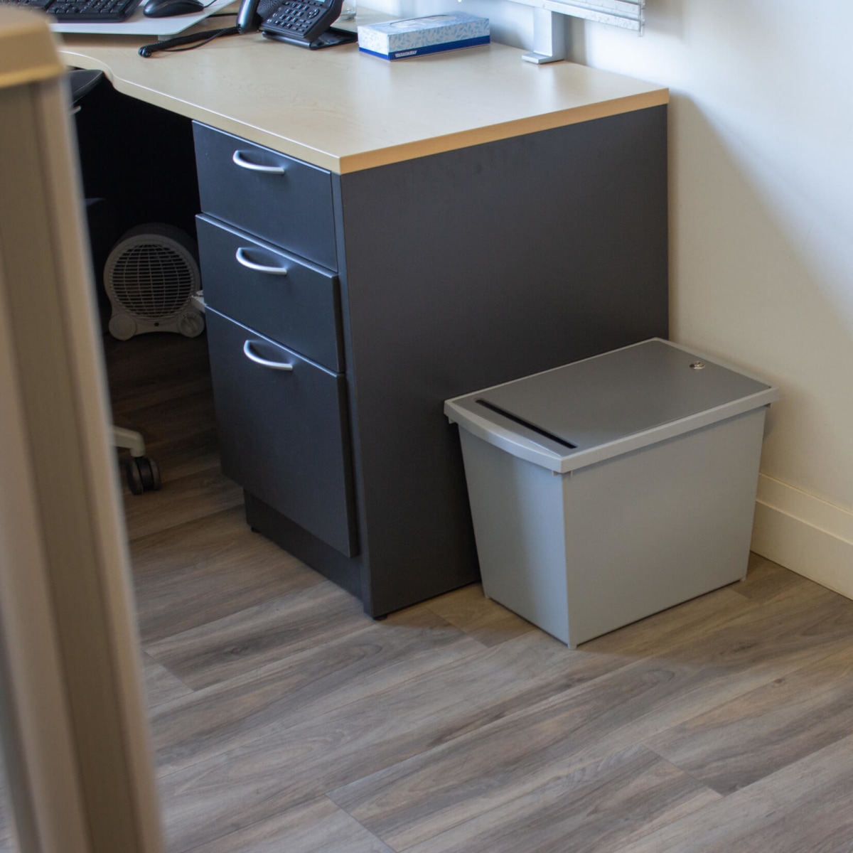 grey plastic personal document container for collecting important papers for shredding in an office sitting on the floor beside a desk