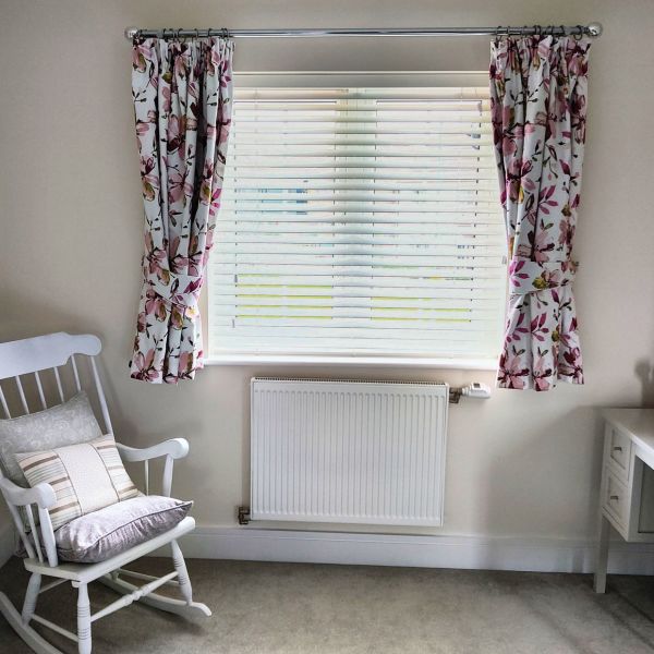 Pink floral pleated curtains, tied back on a silver rod, frame a window; white slatted blinds filter daylight into a pale room with a radiator and a cushioned rocking chair.