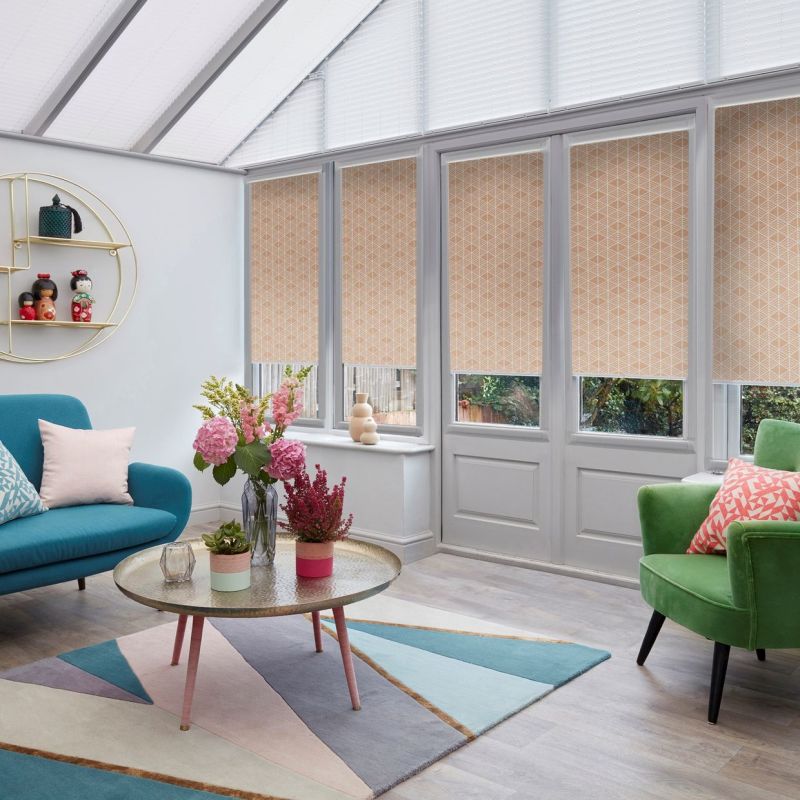 Beige geometric roller blinds on tall conservatory windows, lowered to cover upper panes and filter daylight while revealing lower glass. Sunroom with pastel sofa, armchair, coffee table and garden view.