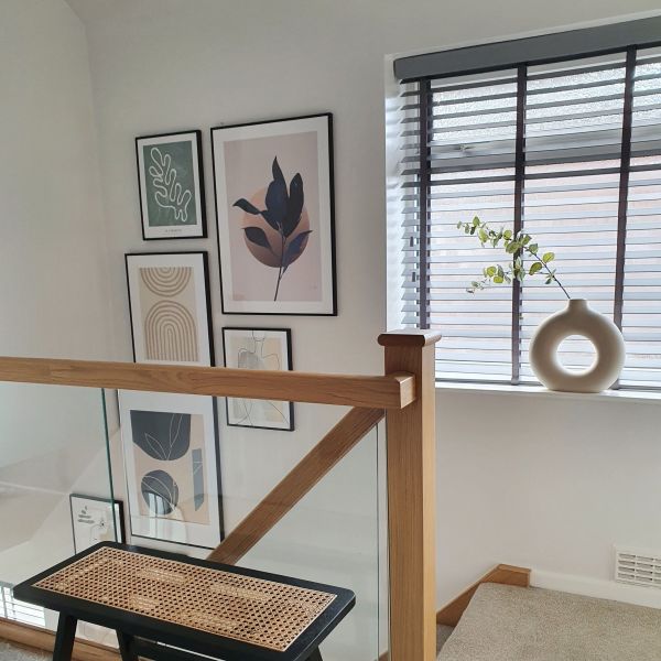 Horizontal slatted blinds on a landing window, partially tilted and filtering light; beside a wood-and-glass stair railing, framed abstract prints on the wall and a round vase on the sill.