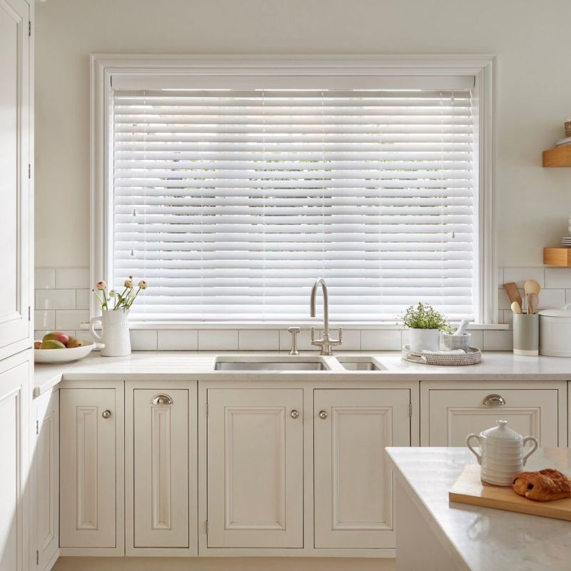 White horizontal Venetian blinds, fully lowered across a wide window with slightly angled slats; filtering soft daylight into a bright, neutral kitchen above a double sink and cream cabinetry.
