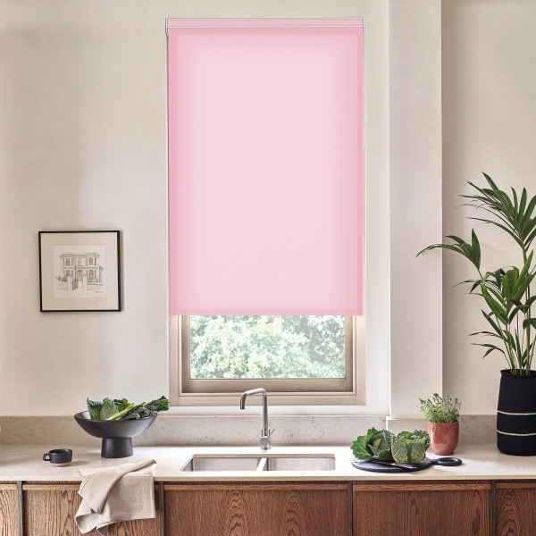 Pink roller blind covers most of a kitchen window, filtering daylight; stainless faucet and double sink below, wood cabinets, countertop with bowls of leafy greens, potted plants, framed sketch.