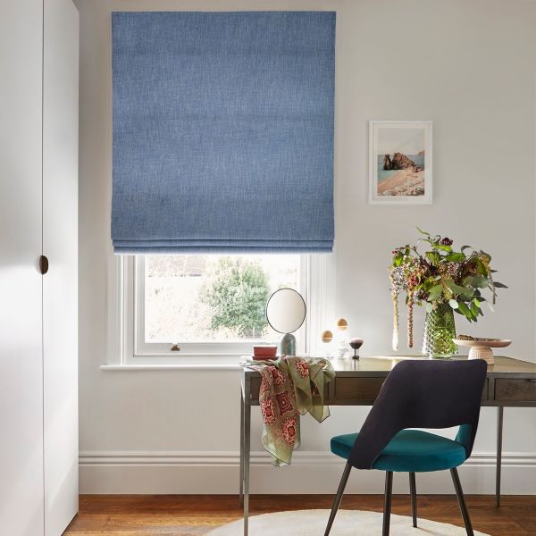 Blue fabric Roman blind lowered over a sash window, textured and folded near sill, filtering daylight; desk with vase of flowers and teal chair below in a bright neutral room.