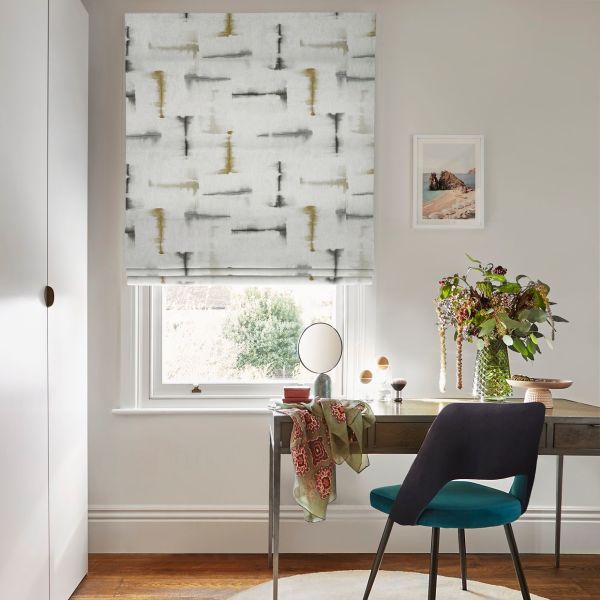 Patterned fabric Roman blind (light gray with gold and charcoal brushstrokes) — partially lowered, filtering daylight — over a desk with teal chair, vase of flowers, pale walls and garden view.